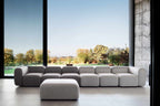 Modular Sofa mit langer Sitzreihe und passendem Hocker vor großer Fensterfront