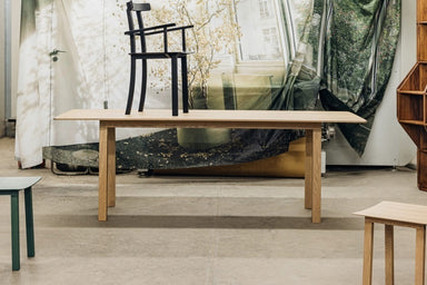 Ein moderner Raum mit einem Holztisch und einem schwarzen Stuhl darauf, daneben ein kleinerer grüner Tisch und im Hintergrund ein großes Fenster mit Vorhängen und einem Blick auf Bäume.